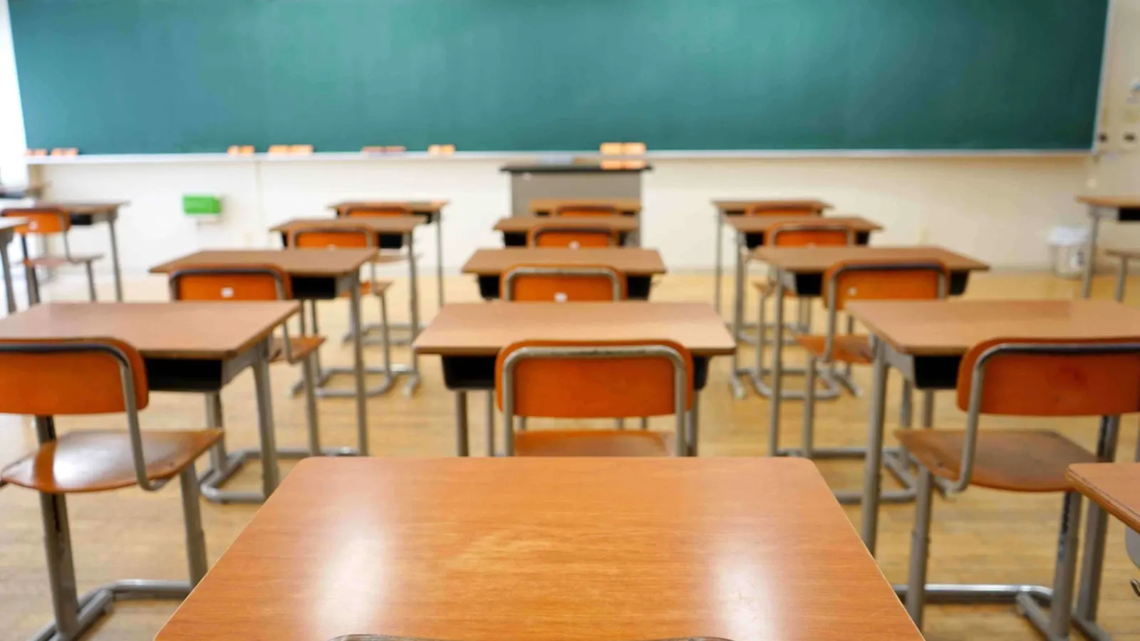 An empty classroom of wooden desks facing a green chalkboard.
