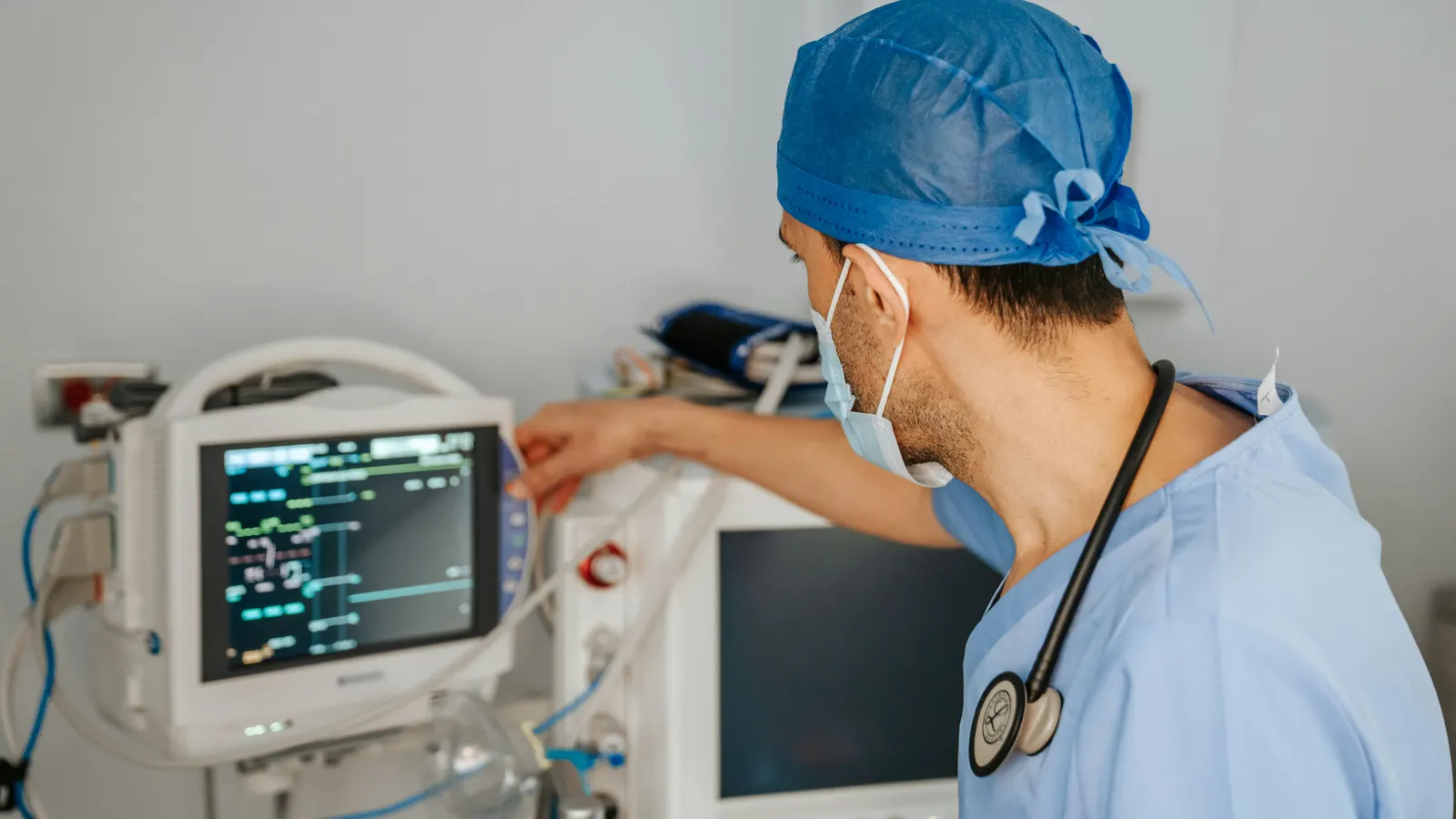 A clinician in scrubs and mask adjusting a bedside patient monitor.